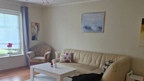 Sala de estar com sofá bege, mesa de centro branca, poltrona branca e ventilador de teto. Há uma janela com vista para o verde e um quadro na parede.