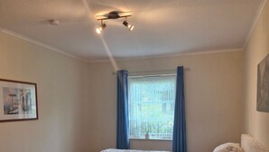 Um quarto com cama, mesa de cabeceira, janela com cortinas, ventilador de teto e um quadro na parede.
