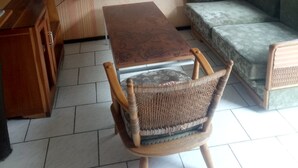 A living room with a wooden table, a wicker chair, a sofa, a television on the wall, and a window with curtains. - Hapo Lodge - Adults Only (Gilgil)