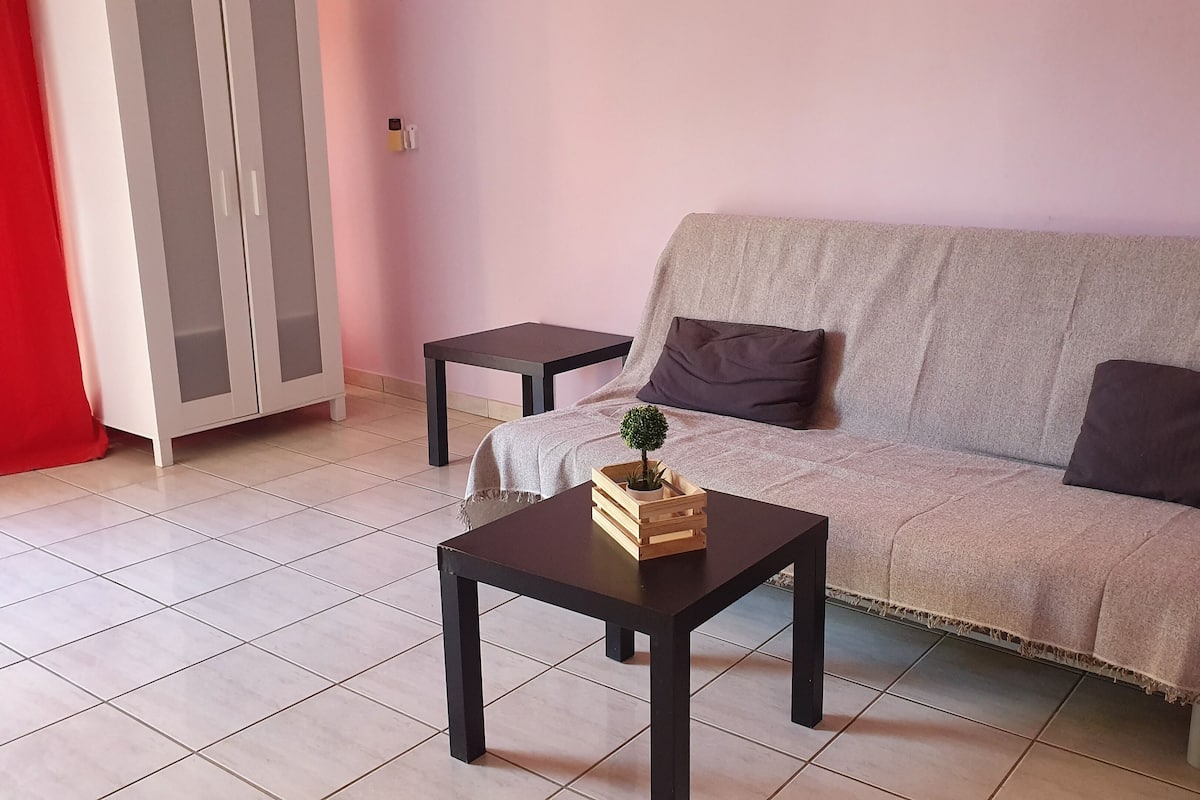 Una sala de estar con un sofá, dos mesitas auxiliares, una pequeña mesa de madera con una planta y una cortina roja.