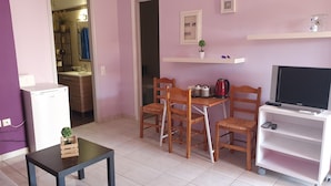 Un espacio habitable compacto con kitchenette, un área de comedor con sillas de madera, una mesa pequeña, un televisor sobre un mueble bajo y un estante con una planta y un reloj.