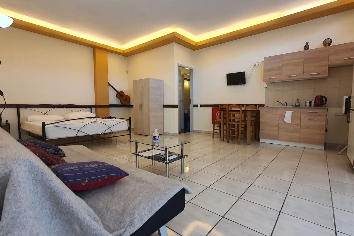 A modern hotel room with a bed, a glass table with a bottle, a guitar, and a kitchen area with wooden cabinets and a refrigerator.