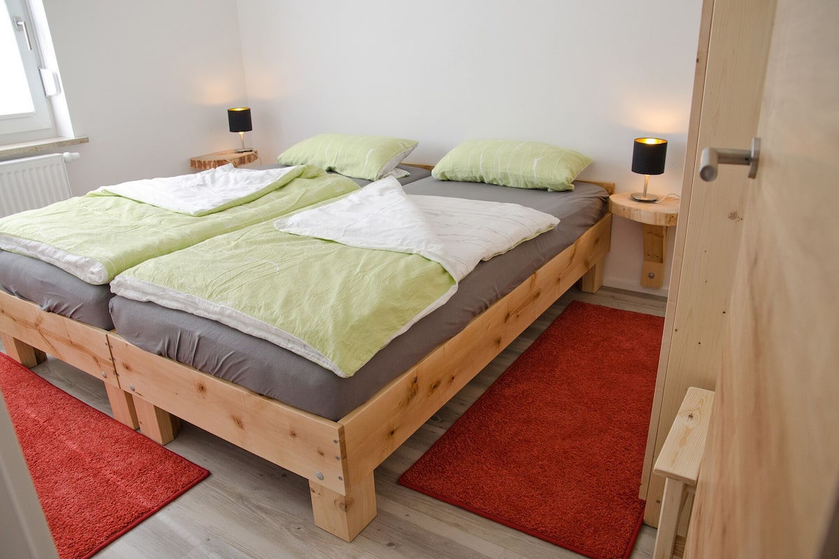 A double bed with green and white bedding, two bedside tables with lamps, a red rug, and a wooden door.