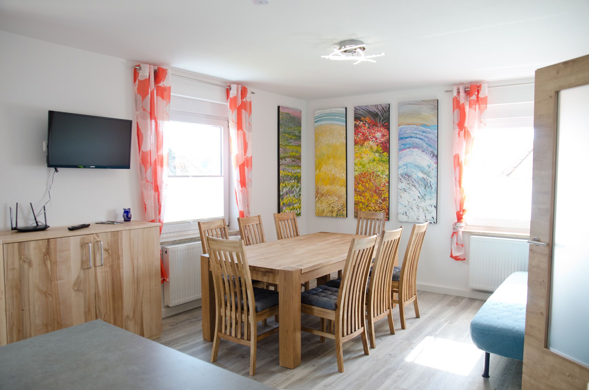 Un comedor con una mesa y sillas de madera, un televisor montado en la pared y obras de arte en las paredes.