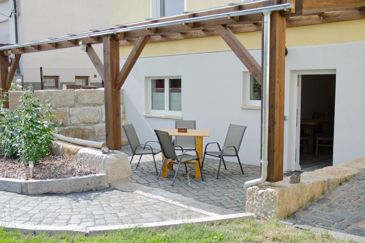 An outdoor patio with a wooden pergola, stone seating, and a small table with chairs.
