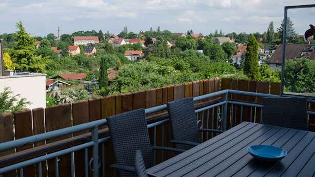 Un balcon avec une table et des chaises, donnant sur un quartier résidentiel avec des maisons et des arbres.