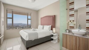 Une chambre moderne avec un grand lit, une vue sur la ville, une coiffeuse avec un lavabo et une tête de lit à motifs.