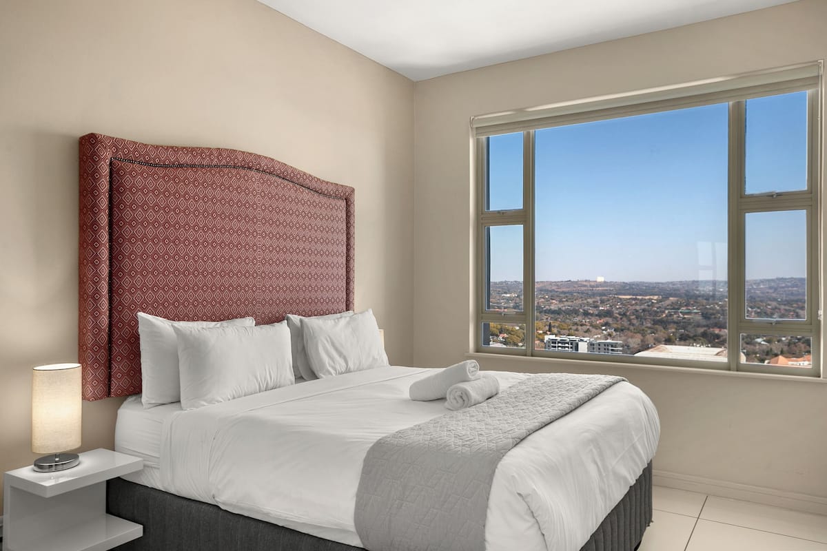 Ein Hotelzimmer mit einem großen Bett, einem Nachttisch mit Lampe und einem Fenster mit Blick auf die Stadt.
