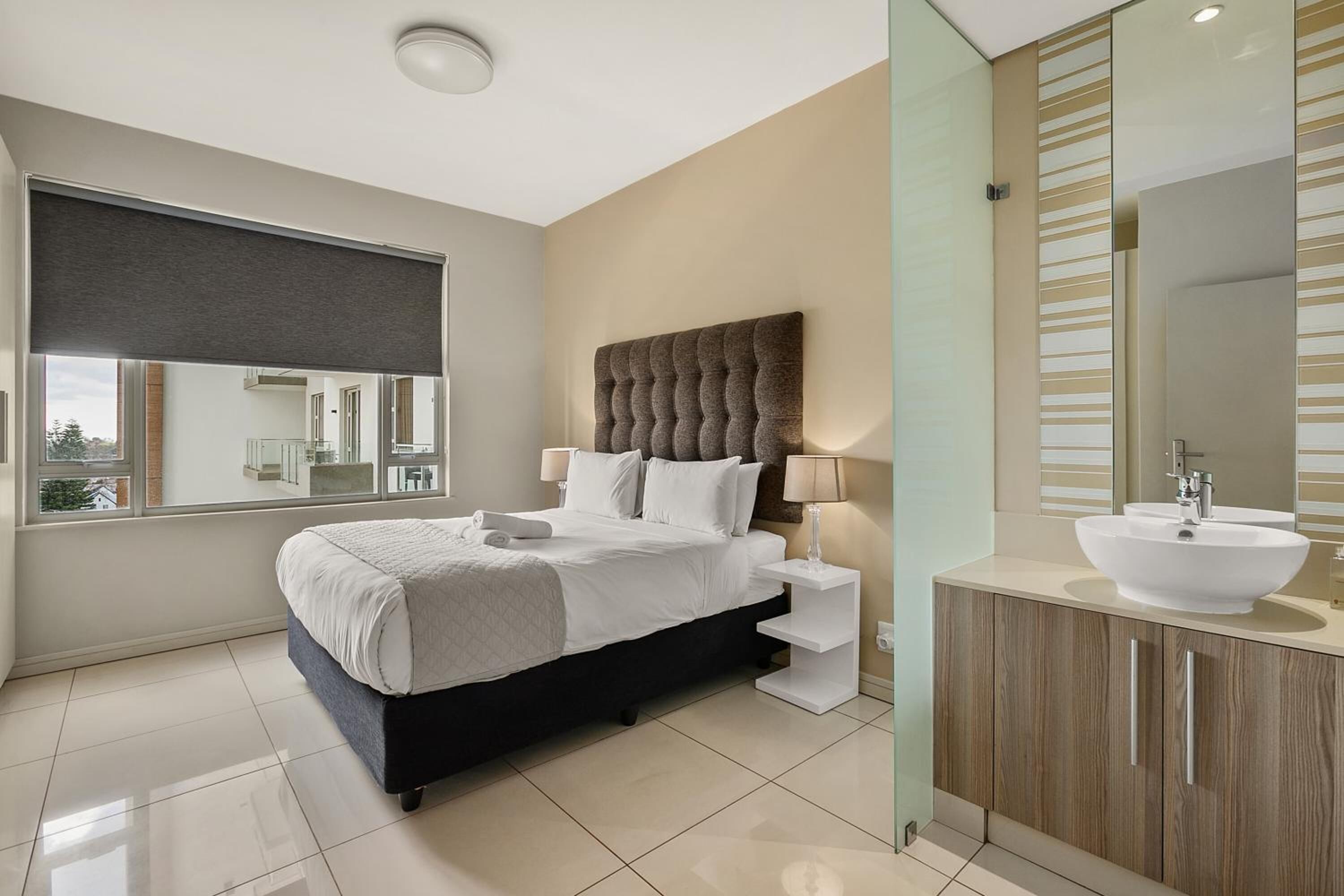 A modern bedroom with a large bed, a bedside table, a large mirror, and a bathroom with a sink and vanity.
