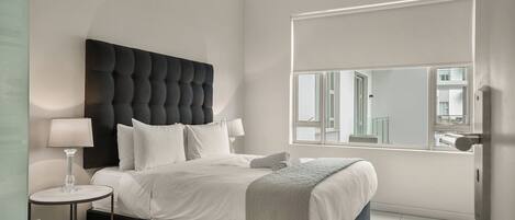 A modern bedroom with a tufted headboard, a round bedside table, a large window with a view, and a neatly made bed.