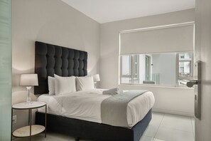 A modern bedroom with a tufted headboard, a round bedside table, a large window with a view, and a neatly made bed. - Easy Stay - The Tyrwhitt (Johannesburg)