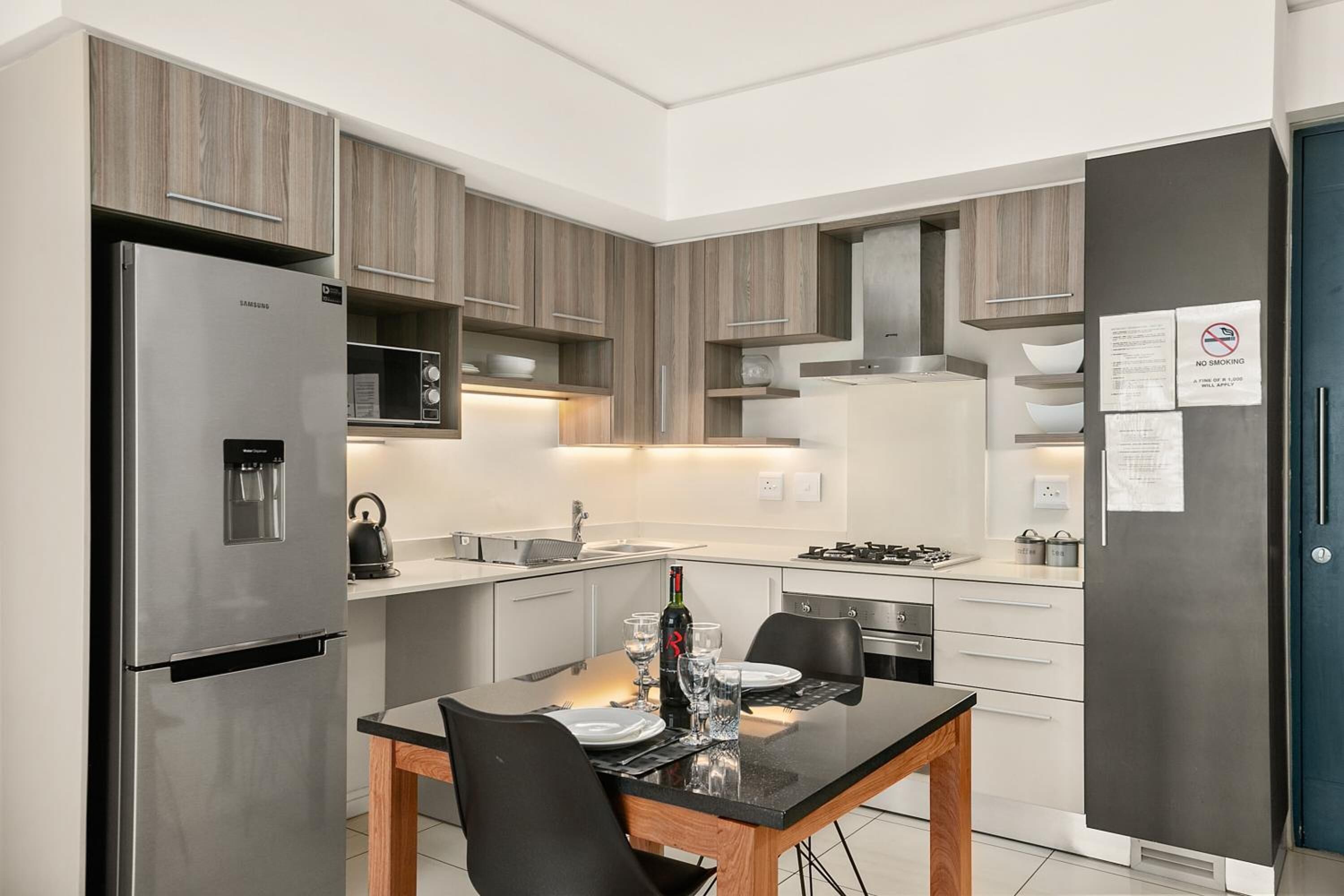 Eine moderne Küche mit Edelstahl-Kühlschrank, Mikrowelle und Herd. Ein Esstisch mit Stühlen und eine Weinflasche sind vorhanden. Die Schränke sind aus hellem Holz, und an der Tür befindet sich ein Rauchverbot-Schild.