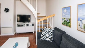 A living room with a grey sofa, a white coffee table, a TV on a white stand, a staircase, and two framed pictures on the wall. - 5 Star Holiday Home in Blavand (Blavand)