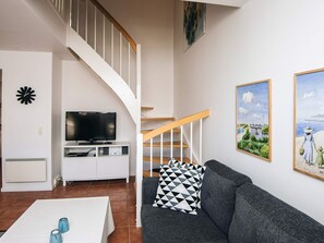A living room with a grey sofa, a white coffee table, a TV on a white stand, a staircase, and two framed pictures on the wall. - 5 Star Holiday Home in Blavand (Blavand)
