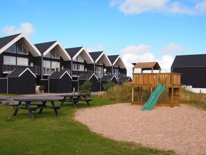 A row of black and white buildings with a sandy area in front, including picnic tables and a wooden play structure with a slide.