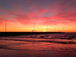A sunset over a beach with waves, a pier, and street lamps. - 5 Star Holiday Home in Lokken (Lokken)