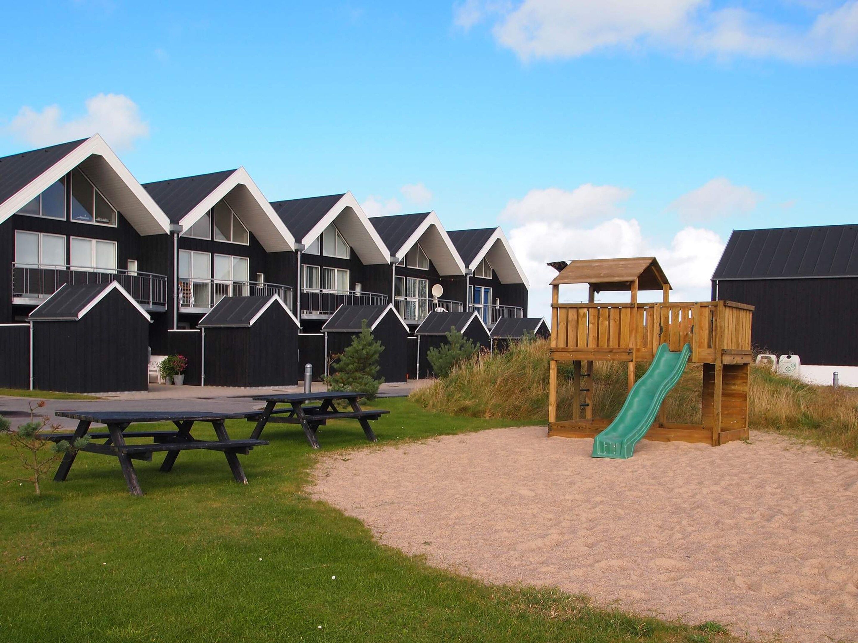 A row of black and white buildings with a sandy area in front, including a wooden play structure with a slide.