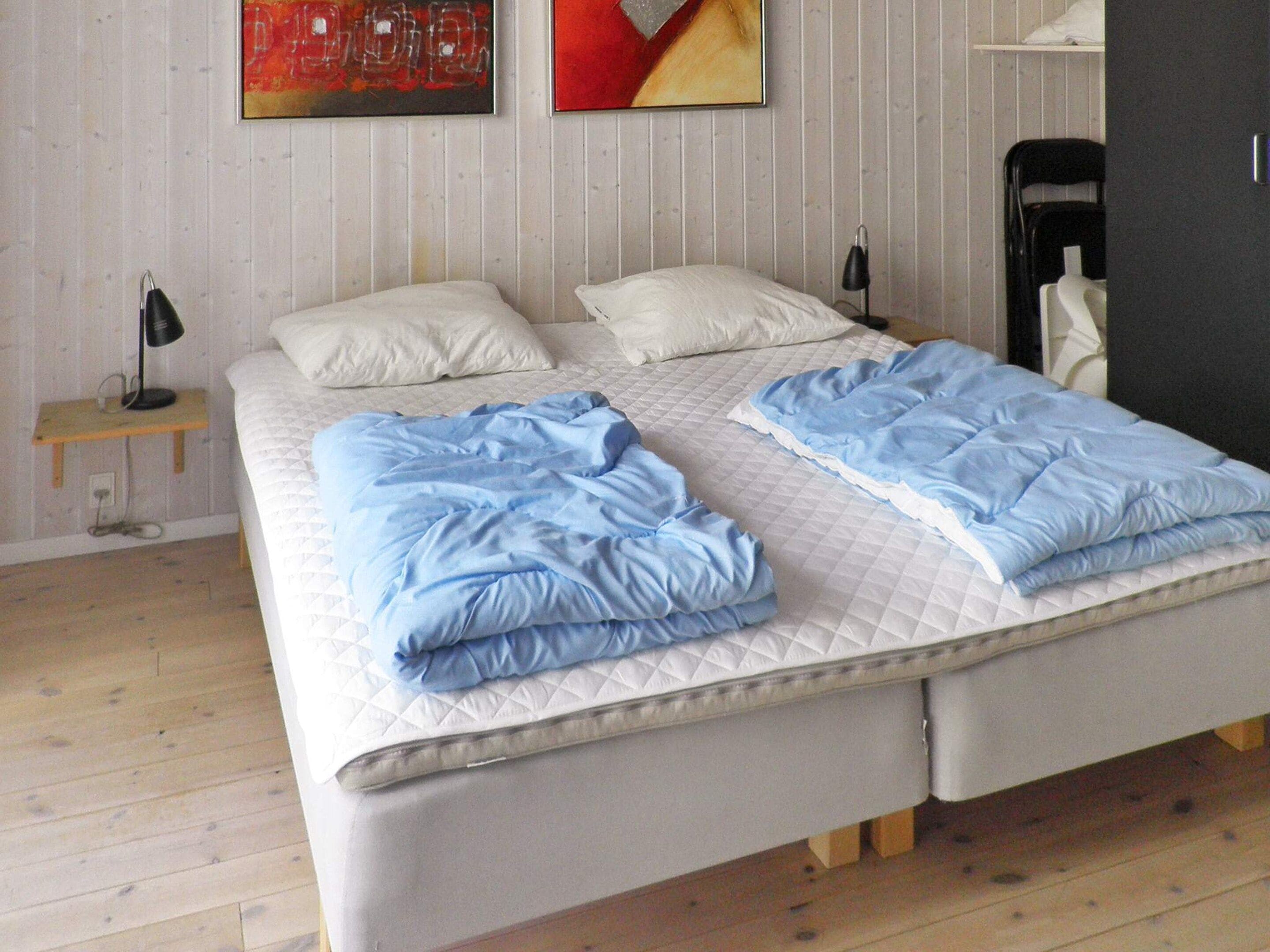 A double bed with a quilted mattress, a bedside table with a lamp, and a chair in a room with wooden flooring and a white wall.