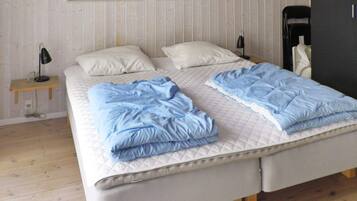 A double bed with a quilted mattress, a bedside table with a lamp, and a chair in a room with wooden flooring and a white wall.