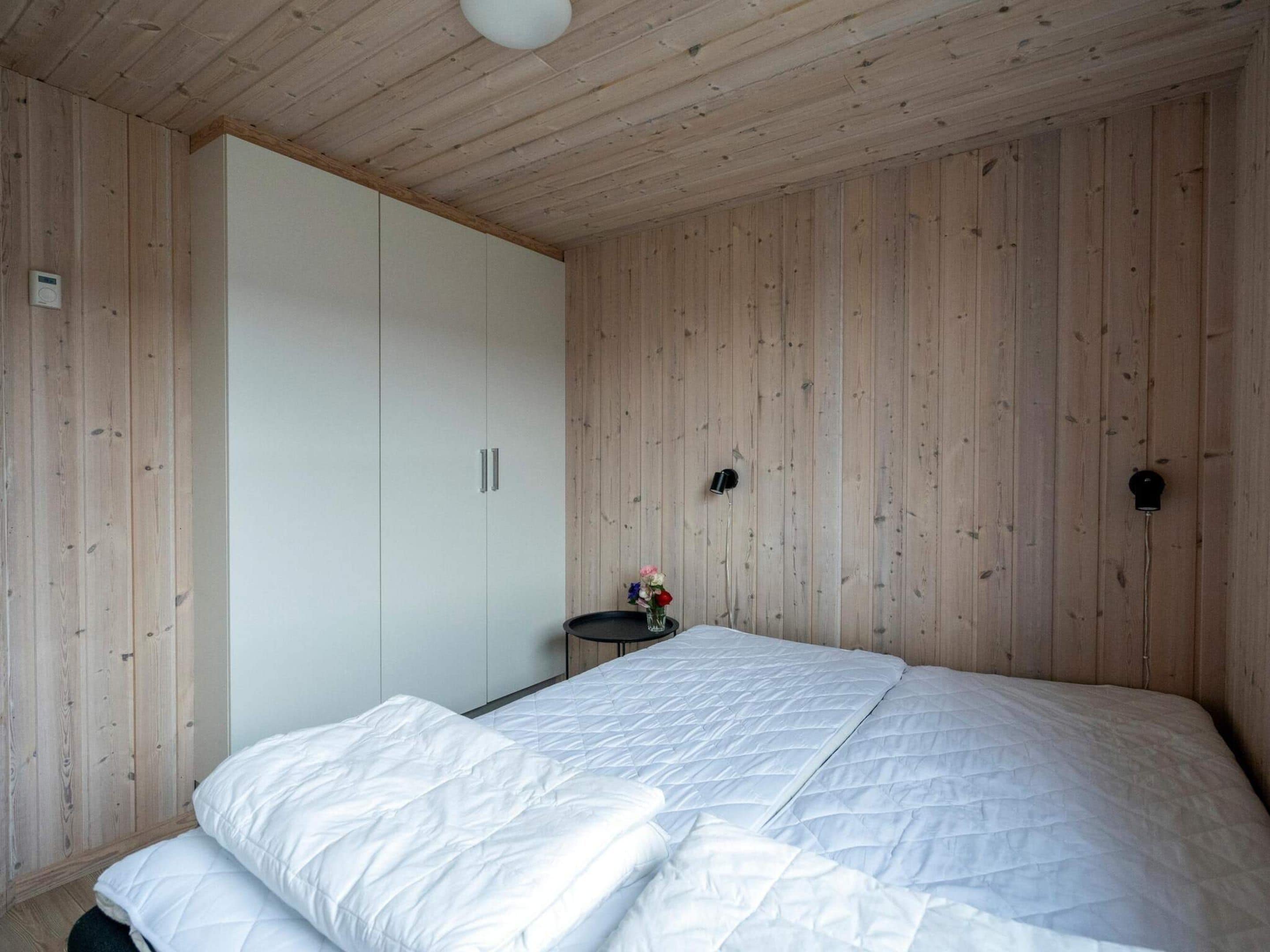 A bedroom with a large bed, wooden walls, a wardrobe, a bedside table with a lamp, and a thermostat.