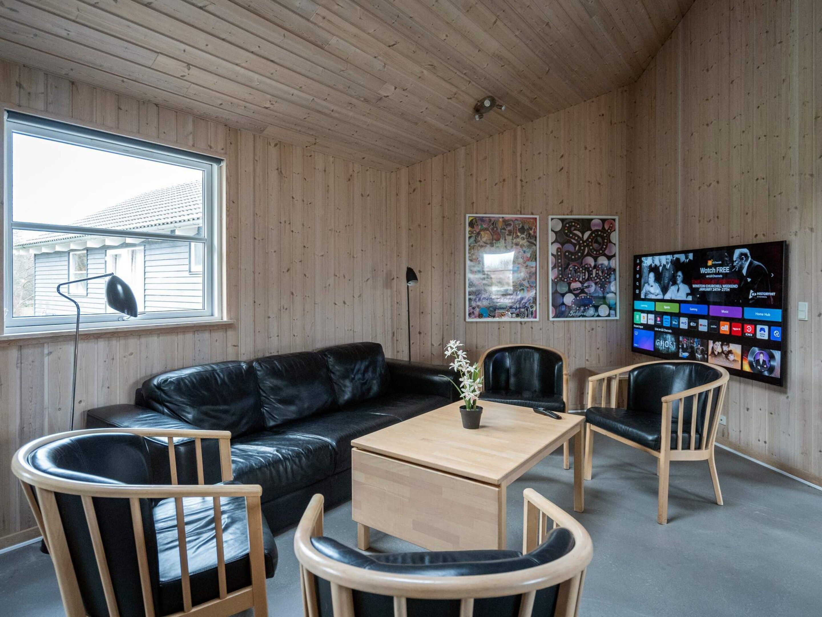 Una habitación con techo de madera, un sofá de cuero negro, sillas de madera, una pequeña mesa de madera y un televisor en la pared mostrando un programa.