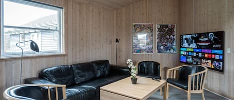 A room with a wooden ceiling, a black leather sofa, wooden chairs, a small wooden table, and a TV on the wall displaying a program.