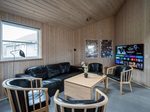 Una habitación con techo de madera, un sofá de cuero negro, sillas de madera, una pequeña mesa de madera y un televisor en la pared mostrando un programa.