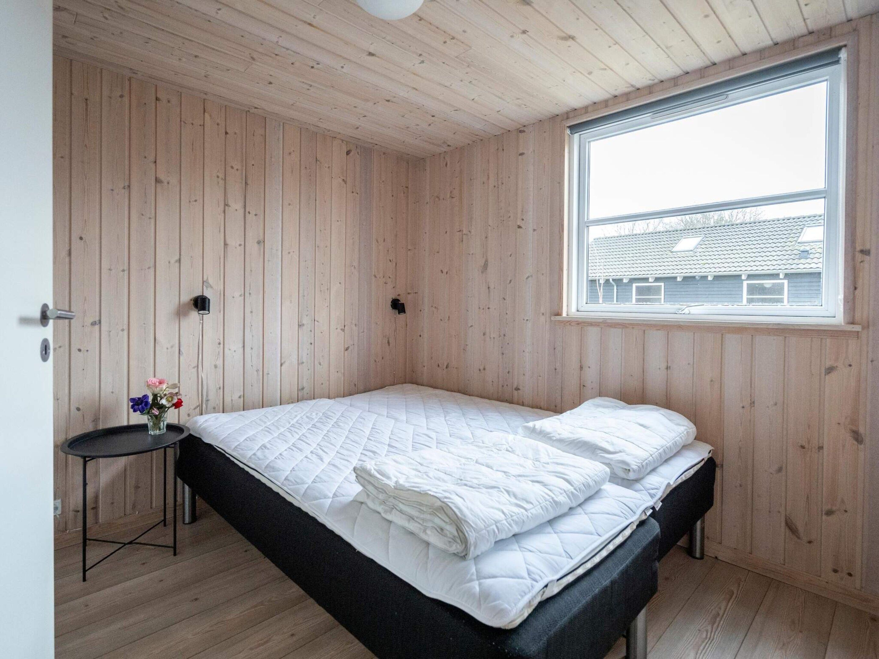 A room with wooden walls, a bed with white bedding, a small table with a vase of flowers, and a window with a view of buildings.