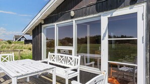 Une terrasse en bois, agrémentée de meubles blancs, dont une table et des chaises, et de grandes portes vitrées donnant sur un espace extérieur.