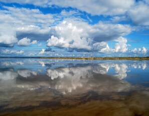 Une étendue d’eau calme où se reflètent les nuages, avec une côte lointaine ponctuée de bâtiments industriels.