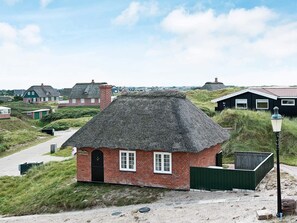 Une maison traditionnelle à toit de chaume, dotée d’un petit porche à l’avant, entourée de dunes herbeuses et d’autres maisons similaires.