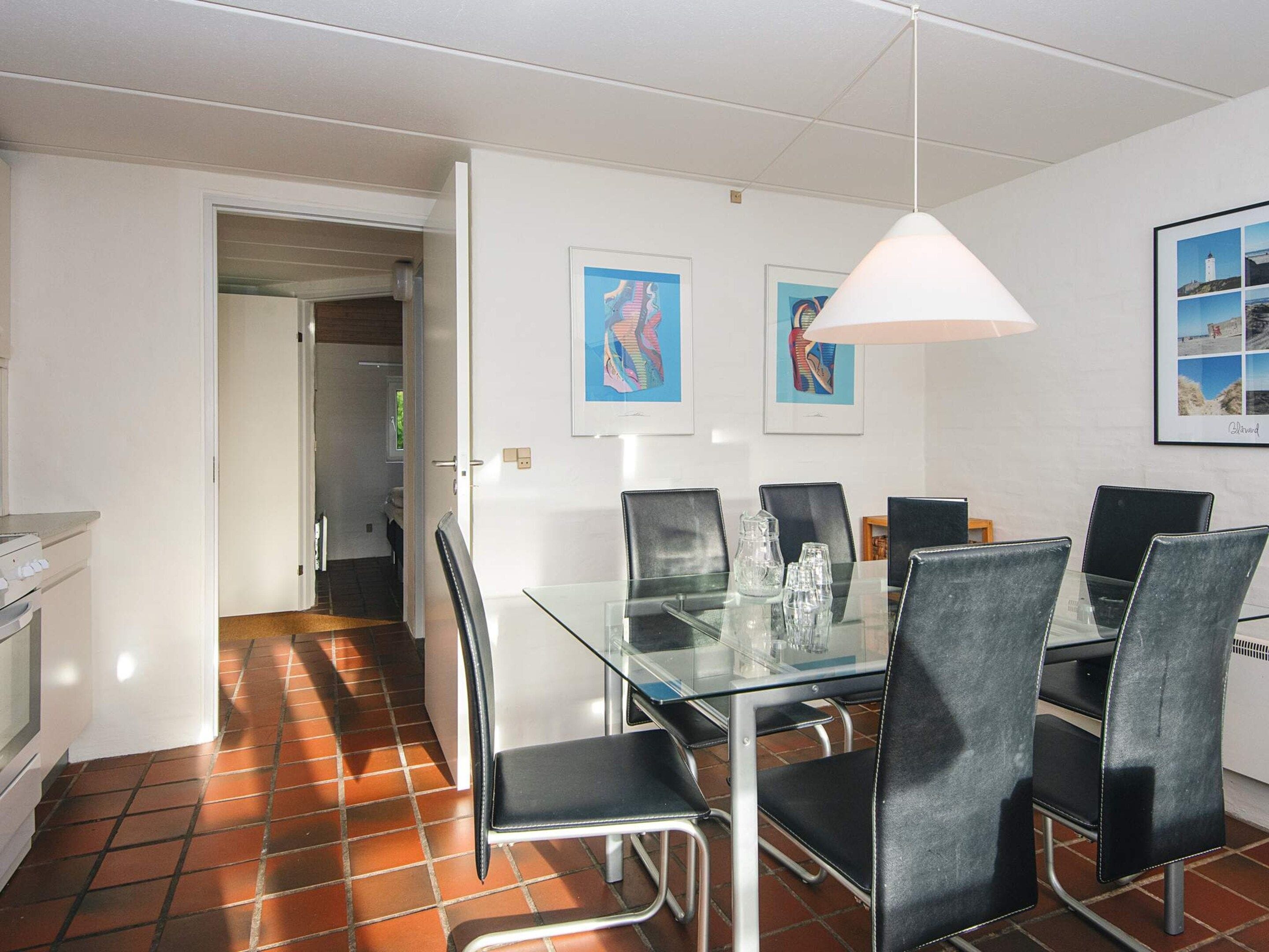 Een eethoek met een glazen tafel, zwarte stoelen, een witte hanglamp en twee kunstwerken in lijsten aan de muur.