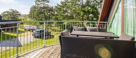 En balkon med en fletsofa og et bord, med udsigt over en parkeringsplads og træer.