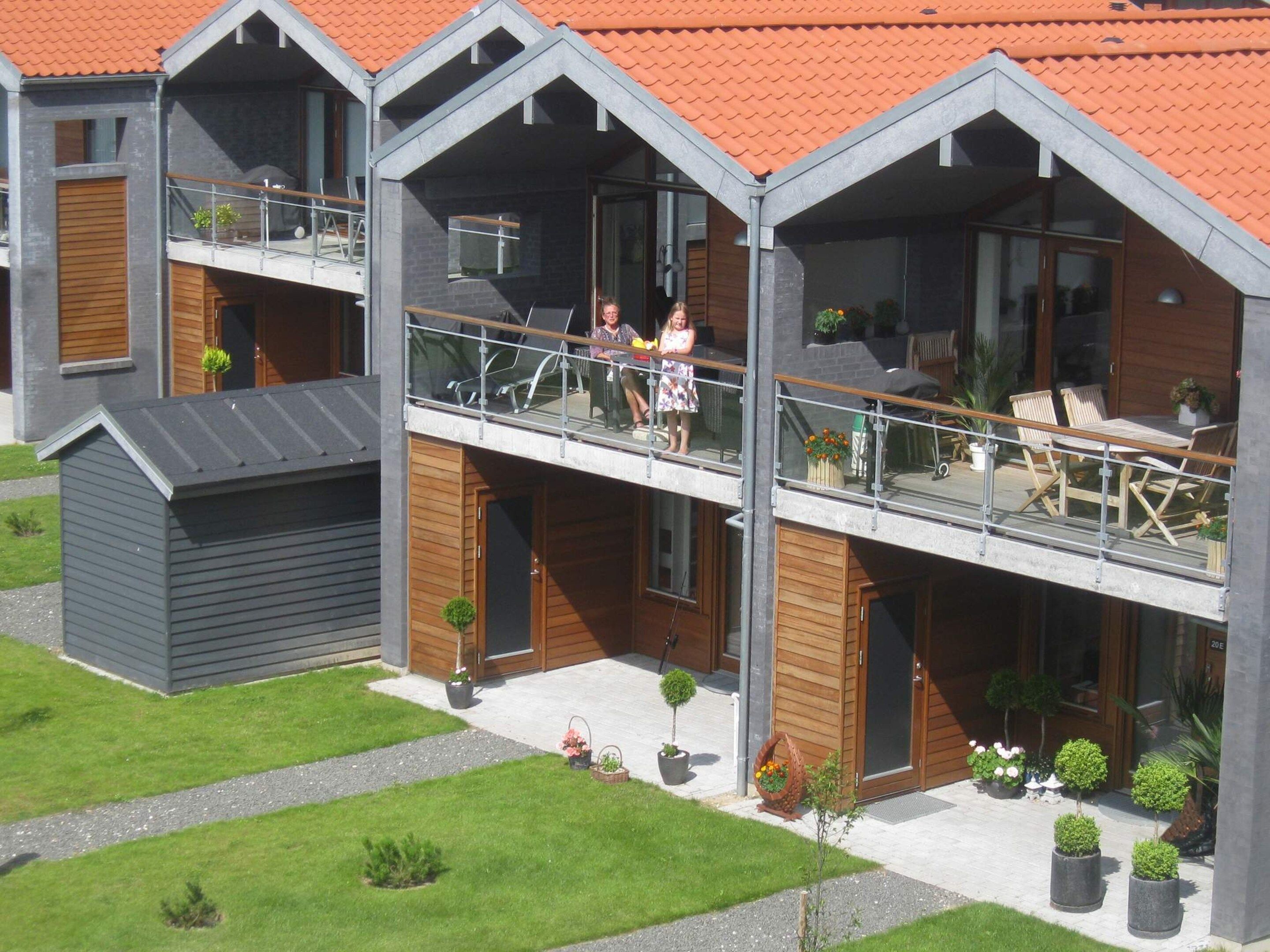 A modern two-story residential building with a balcony, wooden shutters, and potted plants.