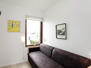 Una habitación con un sofá de color oscuro, una ventana con persianas y dos cuadros en la pared.