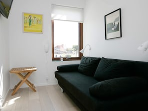 A room with a black sofa, a small wooden table, a window with blinds, and two wall-mounted lamps.
