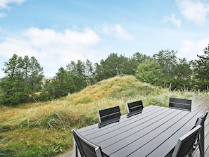 A wooden table with four chairs and two laptops, set outdoors with a grassy hill and trees in the background.