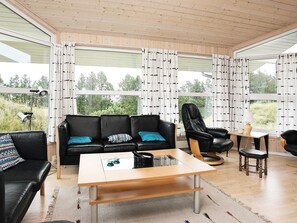 A modern living room with a black leather sofa, a wooden coffee table, and a black leather armchair. Large windows with patterned curtains provide a view of greenery outside.