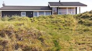 A modern house with a flat roof and large windows, surrounded by grassy dunes.