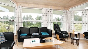 A modern living room with a black leather sofa, a wooden coffee table, and a black leather armchair. Large windows with patterned curtains provide a view of greenery outside.
