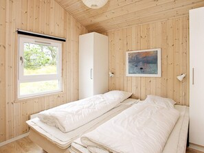A room with wooden walls, a bed with white bedding, a window with a view of greenery, and a painting on the wall.