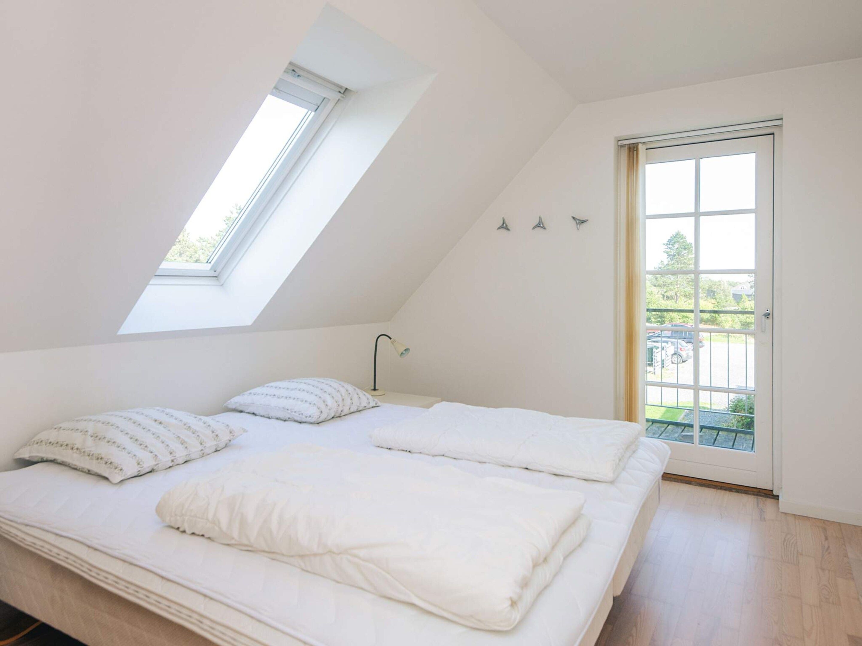 Un dormitorio con techo inclinado, tragaluz, cama con ropa blanca, lámpara de noche y puerta que da a un balcón.