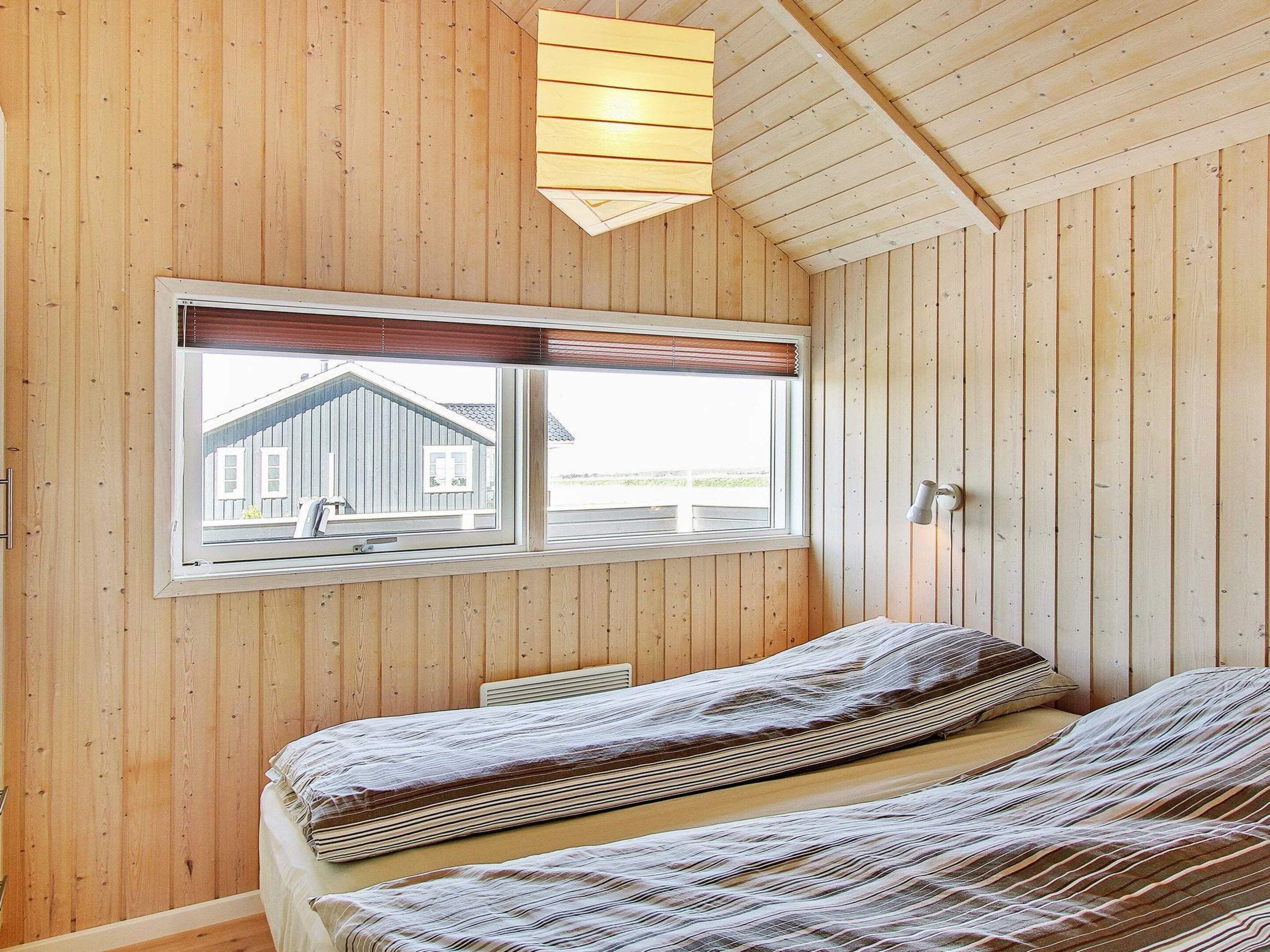 A room with wooden walls, a window with a view of a house, and a bed with striped bedding.