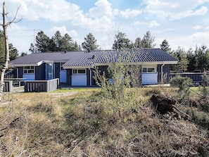 A modern two-story house with a dark roof and light siding, surrounded by a natural landscape with trees and shrubs.