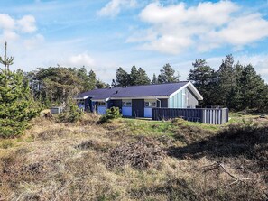 A single-story house with a dark blue exterior and white trim, surrounded by a wooden fence and dense forest.