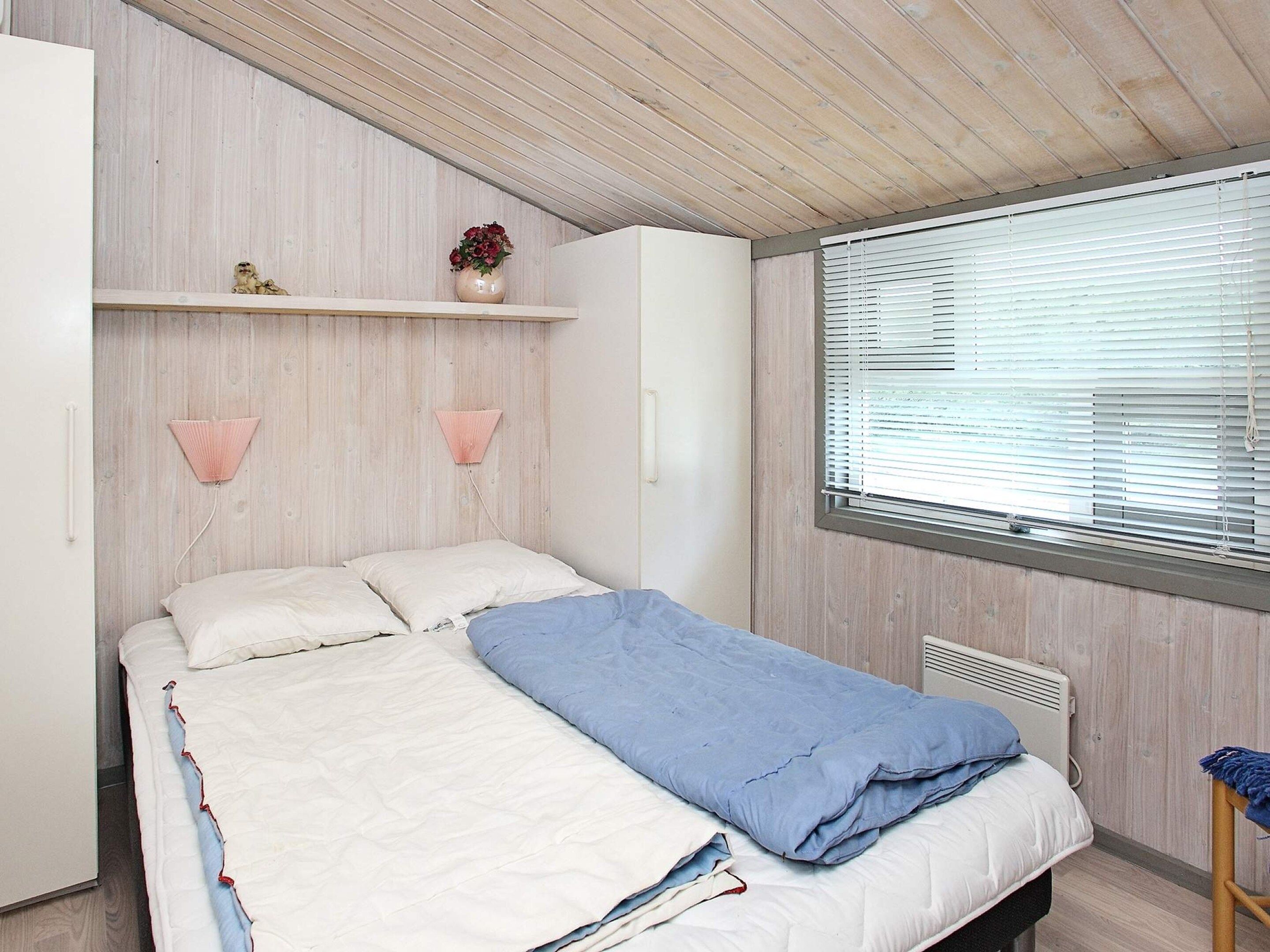 Ein Schlafzimmer mit einem Bett, einem Fenster mit Jalousien, einem Heizkörper und einem Regal mit einer Vase.