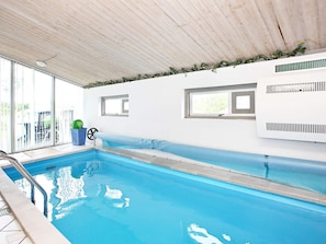 An indoor swimming pool with clear blue water, wooden ceiling, and a bench along the wall.
