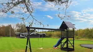 A playground with a swing set and a slide, surrounded by a grassy yard and a building in the background. - 5 Star Holiday Home in Lokken (Lokken)