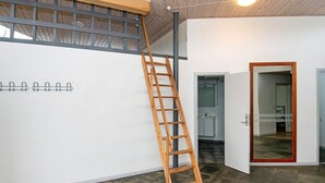 Una habitación con una escalera de madera apoyada en una pared blanca, suelo de baldosas y una puerta que da a otra estancia.