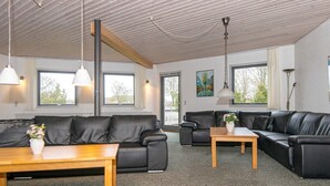 Una sala de estar moderna con dos sofás de cuero negro, una mesa de centro de madera y una mesita con un jarrón. Amplias ventanas ofrecen luz natural y vistas al exterior.